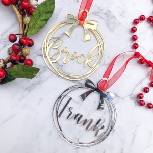 May include: Two personalized Christmas ornaments, one gold and one silver, with the names "Brock" and "Frank" written in a cursive font. Each ornament features a red ribbon bow and a circular design.