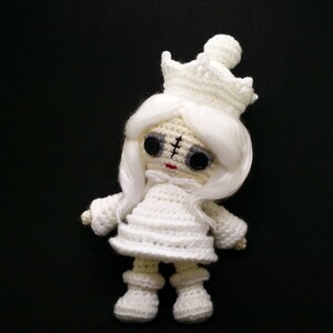 May include: A white crocheted doll wearing a white crown and with long white hair. The doll has black button eyes and a red stitched mouth.