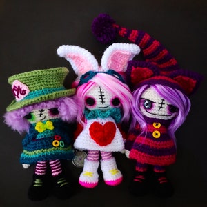 May include: Three crocheted dolls with different outfits and hairstyles. The doll on the left is wearing a green and blue hat and outfit, the doll in the middle is wearing a white dress with a red heart, and the doll on the right is wearing a purple and pink dress with a purple hat.