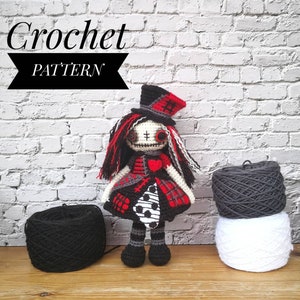 May include: Crochet pattern for a doll with a red, black and white dress, a black and white hat, and black yarn. The doll has a stitched-on face with black eyes and a red heart on its chest. The doll is standing on a wooden surface with two balls of yarn in the background.