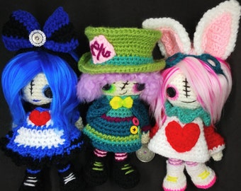 Op de afbeelding: Drie gehaakte poppen met verschillende outfits en kapsels. De pop links heeft blauw haar en een wit-blauwe jurk. De pop in het midden heeft paars haar en een groen-paarse jurk. De pop rechts heeft roze haar en een wit-rode jurk.