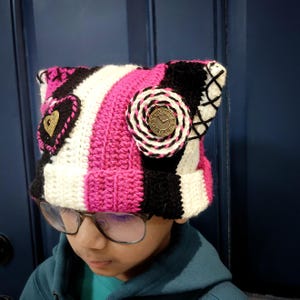 May include: A pink, white, and black striped crocheted hat with a clock and heart embellishments. The hat has cat ears.