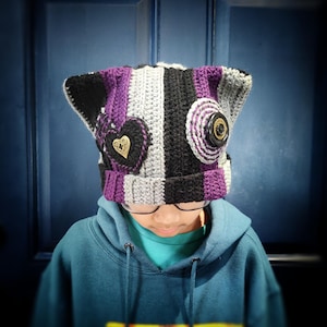 Può includere: Un cappello a uncinetto a righe viola, nero e grigio con orecchie da gatto. Il cappello ha un bottone a forma di cuore e un bottone rotondo con un design a spirale.