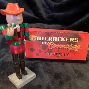 May include: A wooden nutcracker figurine wearing a red hat and a red and green striped outfit. The nutcracker is holding up two fingers and standing next to a red box with the words "Nutcrackers by Commodore Sleep Counselor".