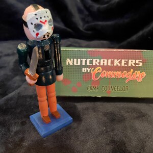 May include: A wooden nutcracker figurine of a character wearing a hockey mask and holding a knife. The figure has orange legs and is standing on a blue base. A green box with the text "NUTCRACKERS by Commodore CAMP COUNSELOR" is behind the figure.