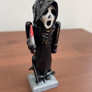May include: A black figurine of the Ghostface character from the Scream horror film series. The figure is holding a silver knife with a red blade. The figure is standing on a gray square base.