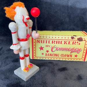 May include: A white and red clown nutcracker with orange hair and a red balloon. The nutcracker is standing on a gray base next to a yellow box with the text "NUTCRACKERS by Commodore DANCING CLOWN".