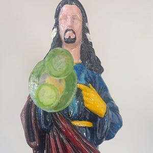May include: A painted statue of a figure with long black hair, a beard, and a blue robe. The figure holds a green object and has a yellow hand pointing towards the chest. The statue has a red cloak and a light skin tone.