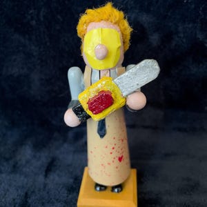 May include: A wooden figurine of a character from a horror movie, holding a chainsaw. The figure has a yellow mask, yellow hair, and a tan apron with red splatters. It stands on a small, square wooden base.