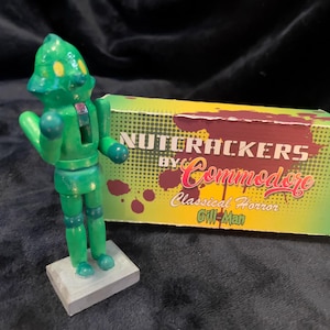May include: A green wooden figure of a robot-like character stands on a gray base next to a box. The box is yellow and green with the words "NUTCRACKERS by Commodore Classical Horror Gill-Man" in red and white.