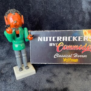 May include: A wooden nutcracker figurine with an orange face, green jacket, and gray pants stands next to a box. The box reads "NUTCRACKERS BY Commodore Classical Horror Wolfman."