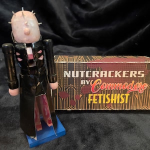 May include: A novelty nutcracker figurine with a pale face and metal pins, wearing a black coat and standing on a blue base. Beside it is a box with the text "NUTCRACKERS by Commodore FETISHIST" in red and gold.
