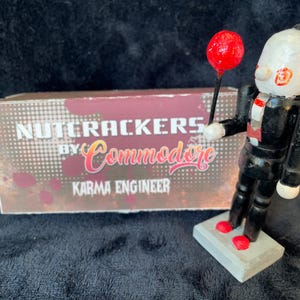 May include: A wooden nutcracker figurine in a black suit, holding a red lollipop. The figurine stands on a gray base with red accents. A box with the text "Nutcrackers by Commodore Karma Engineer" is in the background.