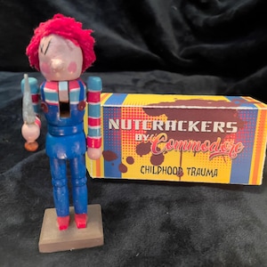 May include: A wooden nutcracker doll with a red wig, blue overalls, and a knife. The doll stands on a small wooden base next to a box that reads "NUTCRACKERS by Commodore CHILDHOOD TRAUMA."