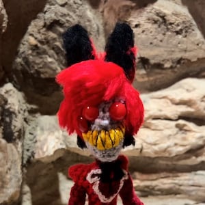 May include: A red and black crocheted doll with a toothy grin and large red eyes. The doll is wearing a red dress with a white heart design and black shoes.