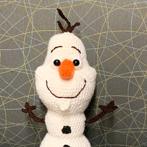 May include: A white crocheted snowman with brown twig arms and a black button nose. The snowman has three black buttons down the front of its body.
