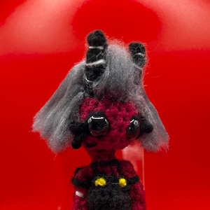 Puede incluir: Una muñeca amigurumi de crochet con cuerpo rojo, pantalones negros y pelo gris. La muñeca tiene ojos amarillos y cuernos negros. La muñeca está de pie sobre una base de plástico transparente.