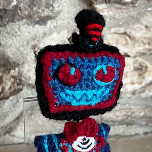 May include: A crocheted plush toy in the shape of a robot with a red and black body, blue arms, and a black top hat with a red stripe. The robot has a red and blue face with a wide, toothy grin.