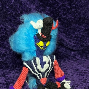 May include: A crocheted plush toy with blue hair, a black top hat, and a red and black striped outfit. The toy is sitting on a purple surface.
