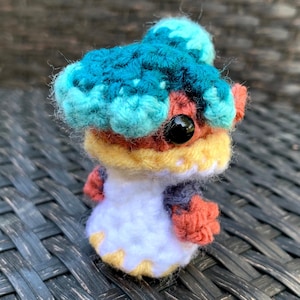 May include: A crocheted plush toy with a blue, yellow and red striped body, a white head and a large blue and turquoise crocheted headpiece. The toy has one black eye.