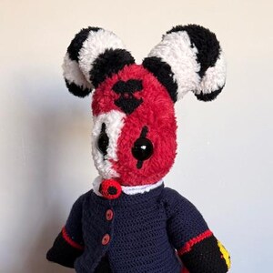 Puede incluir: Una muñeca de peluche hecha a mano con una cara roja y blanca, ojos negros y orejas a rayas. La muñeca lleva una chaqueta azul marino con detalles rojos y pantalones negros. La muñeca está sentada en un otomán azul estampado.