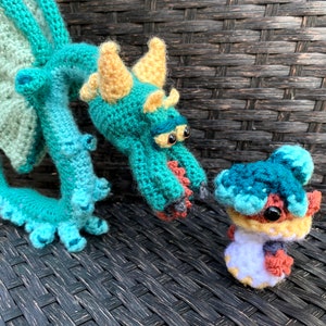 May include: Two crocheted dragon plushies, one large and one small. The larger dragon is teal and yellow with a green wing. The smaller dragon is teal, blue, and orange.