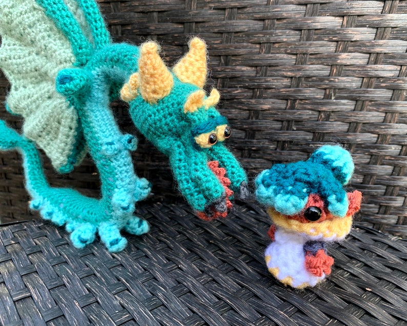 May include: Two crocheted dragon plushies, one large and one small. The larger dragon is teal and yellow with a green wing. The smaller dragon is teal, blue, and orange.