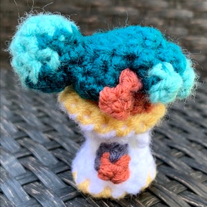 May include: A crocheted amigurumi doll with a teal, blue, and pink body and a yellow and white base.