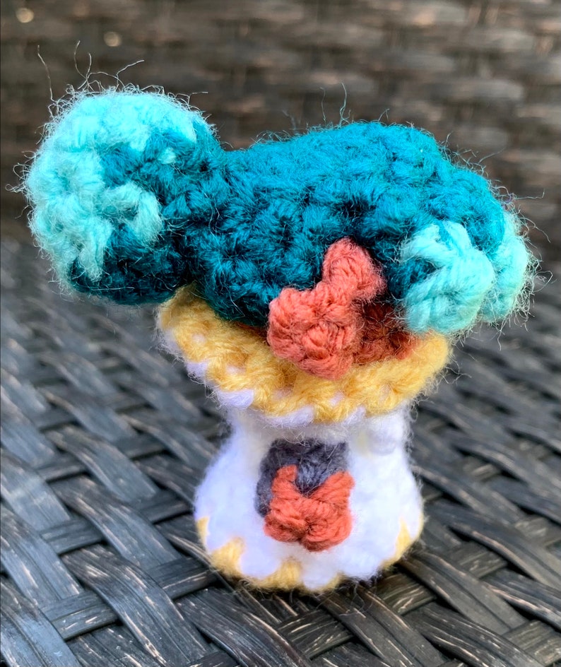 May include: A crocheted amigurumi doll with a teal, blue, and pink body and a yellow and white base.