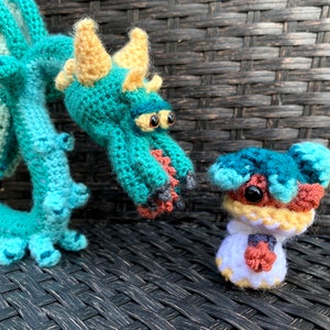 May include: A teal crocheted dragon with yellow horns and a small teal crocheted creature with brown and yellow accents.