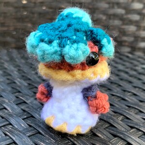 May include: A crocheted amigurumi toy with a blue, teal, and yellow head, red and yellow body, and white dress. The toy has a black eye and is sitting on a woven surface.