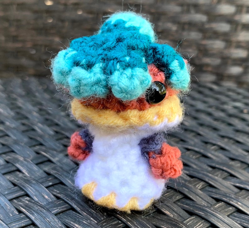 May include: A crocheted amigurumi toy with a blue, teal, and yellow head, red and yellow body, and white dress. The toy has a black eye and is sitting on a woven surface.