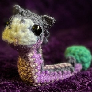 May include: A crocheted plush toy shaped like a worm with a grey and white head, purple and grey body, and a green tail. The toy is lying on a purple fuzzy surface.