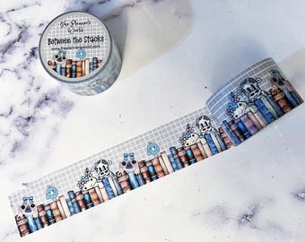May include: Decorative washi tape roll with a repeating pattern of cartoon cows, books, and birds. The tape is gray with a grid pattern and the text "Between the Stacks" is printed on the roll. The Planner's World website is also printed on the roll.