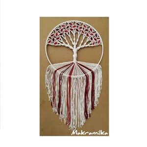 Puede incluir: Un tapiz de macramé blanco en forma de árbol con hojas rojas. El árbol está rodeado de un círculo blanco y tiene una larga franja de hilo blanco y rojo.