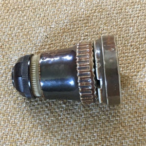 Puede incluir: Un portalámparas vintage marrón y beige con un botón negro. El portalámparas tiene un diseño acanalado y una base de metal. El objeto está sobre una superficie de tela beige.