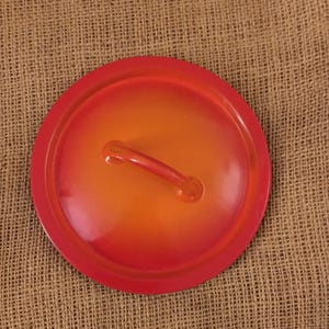 May include: A round, red and orange enameled pot lid with a handle. The lid has a gradient color scheme, transitioning from red to orange. The handle is a matching orange color.