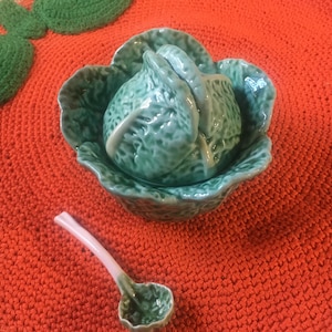 May include: A ceramic cabbage-shaped serving dish with a matching ladle. The dish is a glossy teal green, and the ladle has a pink handle. The set sits on a bright orange crocheted placemat, with green crocheted leaves.