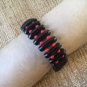 May include: A black bracelet with a glossy finish, featuring a series of curved, oblong segments. Each segment is connected by small, round red beads, creating a striking contrast. The bracelet is displayed on a neutral-toned surface.