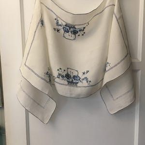 May include: Cream-colored linen tablecloth with blue floral embroidery. The embroidery features a basket of flowers and a vase of flowers. The edges are finished with a blue stitched border. The tablecloth is hanging on a wooden hanger.