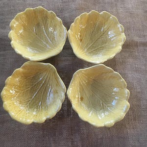 May include: Four yellow ceramic leaf-shaped bowls. Each bowl has a detailed leaf vein pattern and a scalloped edge. The bowls are arranged in a square formation on a textured, neutral-toned surface.