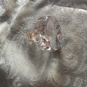 May include: Clear glass bottle stopper with a textured, faceted design. The stopper has a round top and a short, cylindrical neck.