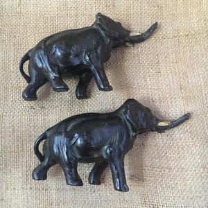 May include: Two black elephant figurines with ivory-colored tusks are displayed on a textured, tan surface. The elephants are in a walking pose, showcasing detailed features and a vintage aesthetic. The figurines are likely decorative items.