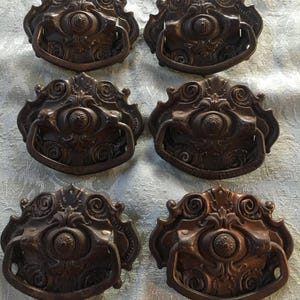 May include: Six antique bronze drawer pulls with ornate, scrollwork designs. Each pull features a central, oval-shaped element with a handle below. The pulls are arranged in two rows of three, showcasing their intricate details and aged patina.