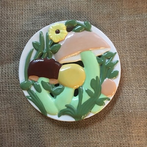 May include: A white ceramic trivet with a mushroom design. The trivet features three mushrooms with green leaves and a yellow flower.