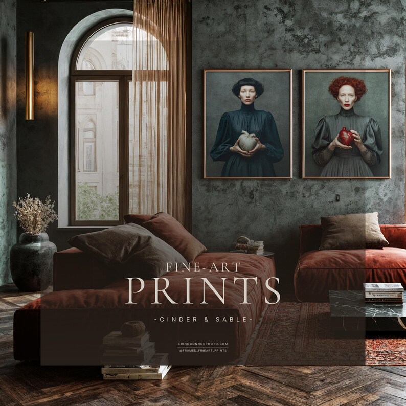 Fine art print of woman with red hair holding a red anatomical heart framed in light wood and displayed beside companion piece in moody living room with rust velvet sofas, arched window, and dark textured walls.
