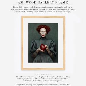 Cinder fine art print framed in a light ash wood gallery frame with white matte, featuring a red-haired woman in a dark dress holding a heart-shaped vessel on a deep green backdrop.