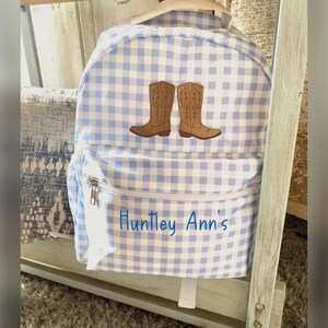 Puede incluir: Una mochila a cuadros azules y blancos con un bolsillo frontal y cierre de cremallera. La mochila presenta un dise&ntilde;o bordado de dos botas de vaquero marrones. El nombre "Huntley Ann's" est&aacute; bordado en el bolsillo delantero.