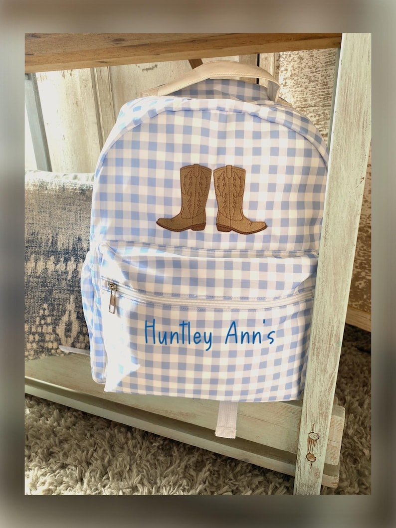 Puede incluir: Una mochila a cuadros azules y blancos con un bolsillo frontal y cierre de cremallera. La mochila presenta un dise&ntilde;o bordado de dos botas de vaquero marrones. El nombre "Huntley Ann's" est&aacute; bordado en el bolsillo delantero.