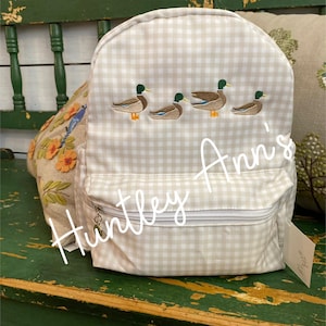 May include: A beige and white gingham backpack with embroidered ducks. The backpack has a zippered pocket and a small tag that says "Huntley Ann's".